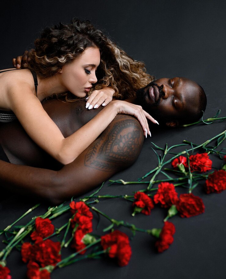 Foto grátis cortada de mulher apaixonada com penteado afro vestindo sutiã deitado no homem afro-americano tatuado bonito nu com os olhos fechados perto de cravos vermelhos dispersos