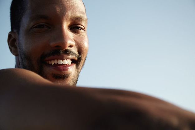 Foto grátis cortada o retrato do jovem africano alegre com barba pequena sentado na calçada, aproveitando o tempo ensolarado, sorrindo