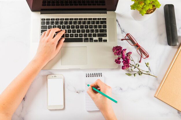 Cortar as mãos usando laptop e fazendo anotações na mesa de mármore