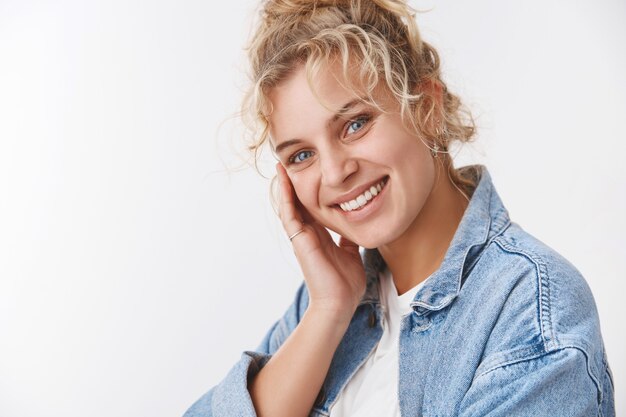 Cosmetologia, conceito de bem-estar. Atraente garota européia de cabelos louros sorrindo corada fofa cabeça inclinada tocando a pele limpa e perfeita como o rosto depois de aplicar o produto para a pele, sorrindo satisfeita