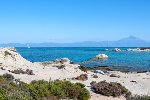 Costa do mar Egeu com pessoas que nadam, rochas sobre a água e terra com barco à distância, vegetação em primeiro plano, água azul, Grécia