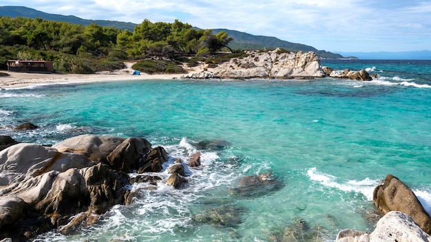 Costa do mar Egeu com vegetação ao redor, rochas, arbustos e árvores, água azul com ondas, Grécia
