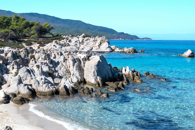 Costa do mar Egeu com vegetação ao redor, rochas e arbustos, água azul e pessoas em repouso, Grécia