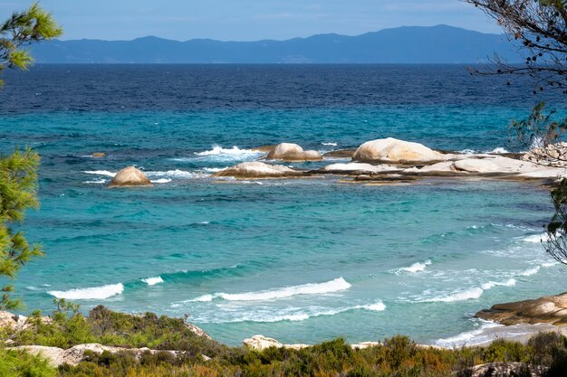 Costa do mar Egeu com vegetação ao redor, rochas e árvores, água azul com ondas, Grécia