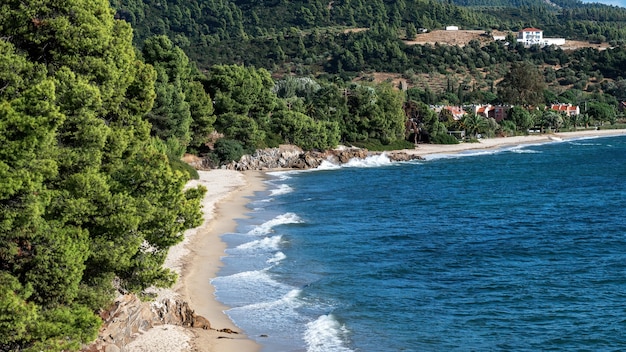 Costa do mar Egeu da Grécia, colinas rochosas com árvores e arbustos em crescimento, praia com ondas, edifícios localizados perto da costa
