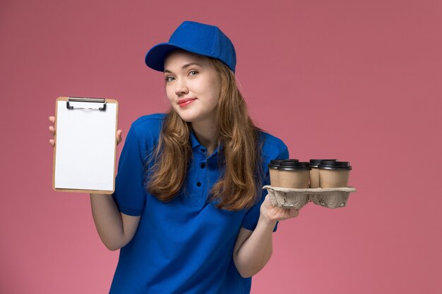 Courier feminina de uniforme azul segurando um bloco de notas e entregando xícaras de café na empresa de uniforme rosa claro