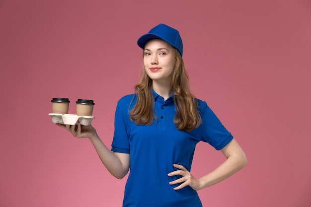 Courier feminina de uniforme azul segurando xícaras de café marrons e posando na mesa rosa da empresa de entrega de uniforme