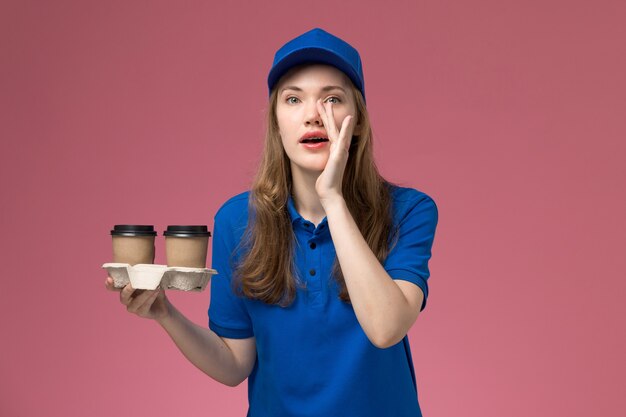 Courier feminina de uniforme azul segurando xícaras de café marrons sussurrando na mesa de trabalho rosa