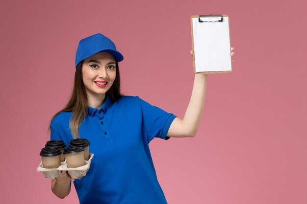 Courier feminina de vista frontal com uniforme azul e capa segurando xícaras de café e um bloco de notas sorrindo na parede rosa