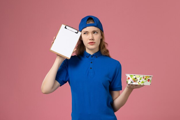 Courier feminino com capa uniforme azul segurando o bloco de notas e a tigela de entrega na parede rosa, trabalhador de entrega de serviço.