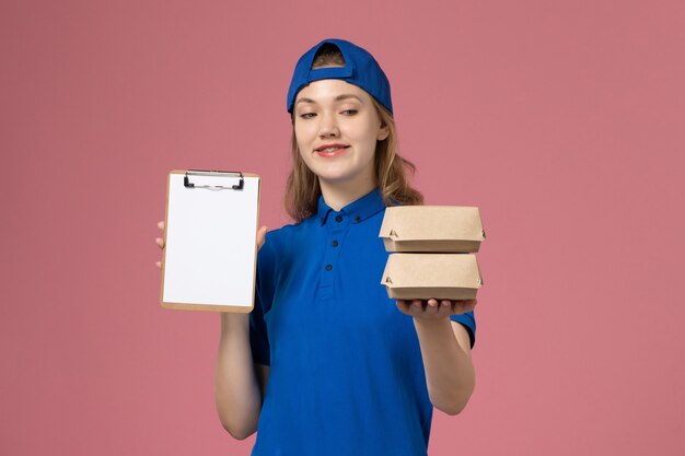 Courier feminino com capa uniforme azul segurando pequenos pacotes de comida para entrega e um bloco de notas no fundo rosa. Trabalhador de funcionário de serviço de entrega