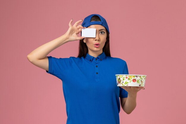 Courier feminino de capa uniforme azul segurando a tigela de entrega com cartão na parede rosa claro, entrega de funcionário de trabalho de serviço