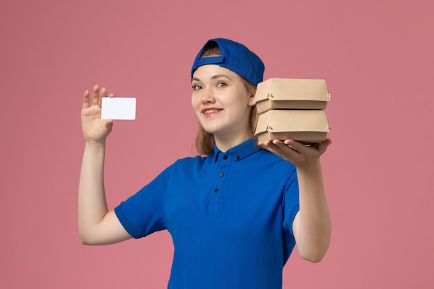 Foto grátis courier feminino de frente para a capa uniforme azul segurando pequenos pacotes de comida para entrega e um cartão no fundo rosa.