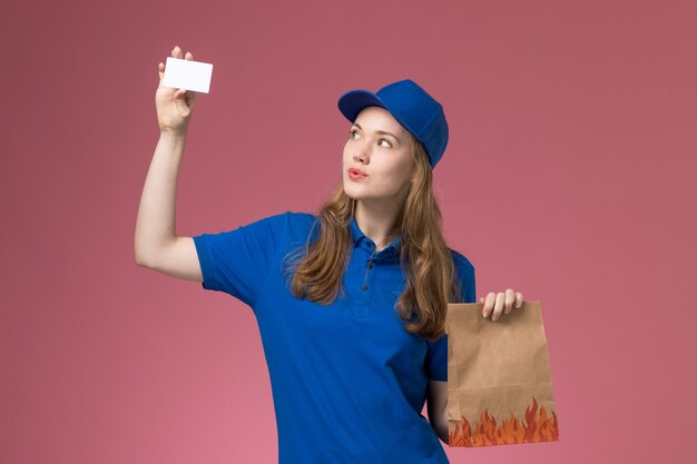 Courier feminino de uniforme azul segurando um cartão branco e um pacote de comida na mesa rosa. Uniforme de serviço.