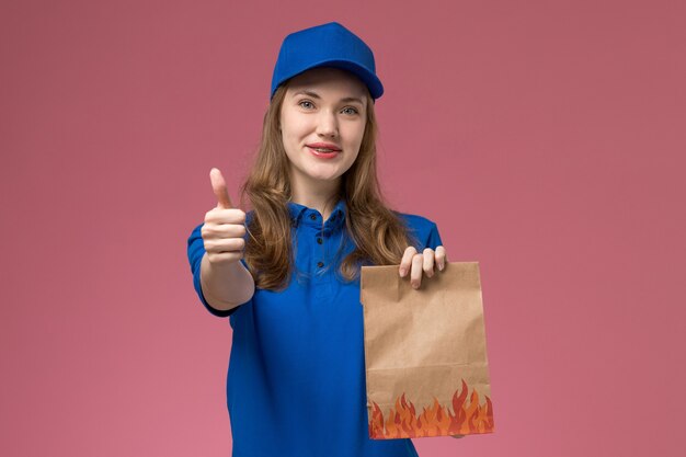 Courier feminino de uniforme azul segurando um pacote de comida mostrando como sinal na mesa rosa trabalho trabalhador serviço uniforme empresa de vista frontal