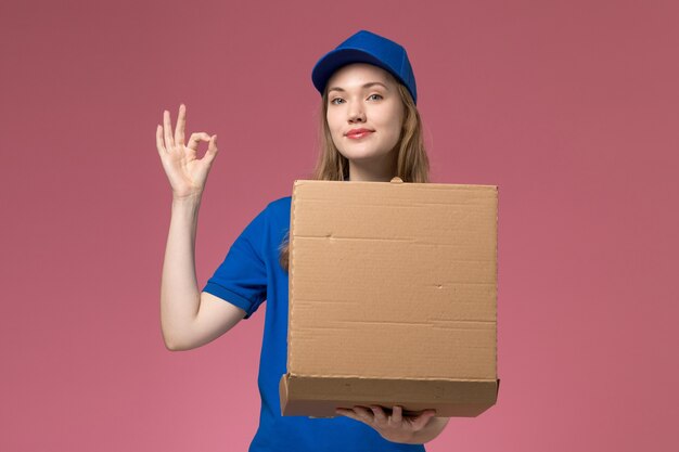 Courier feminino de uniforme azul segurando uma caixa de entrega de comida sorrindo no fundo rosa.