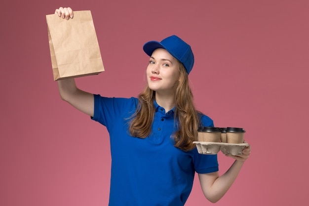 Courier feminino de uniforme azul segurando xícaras de café marrom com pacote de comida sorrindo na mesa rosa uniforme de serviço.