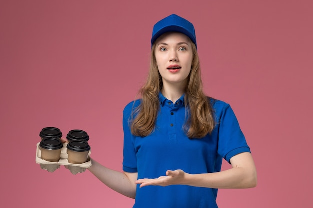 Courier feminino de uniforme azul segurando xícaras de café marrom no fundo rosa uniforme de serviço da empresa.