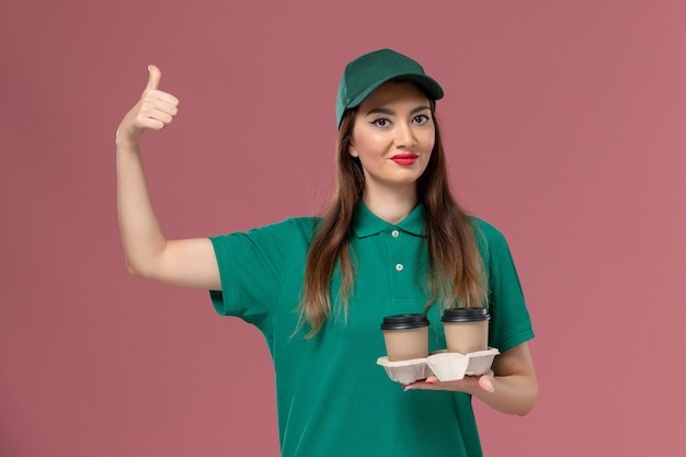 Courier feminino de uniforme verde e capa segurando xícaras de café de entrega na mesa rosa claro.
