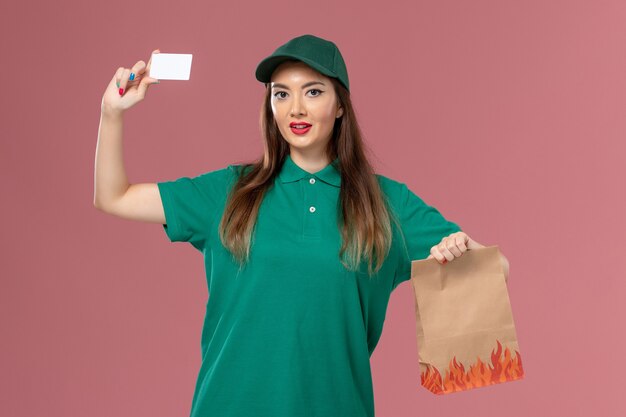 Courier feminino de uniforme verde segurando um cartão branco e um pacote de comida na parede rosa.