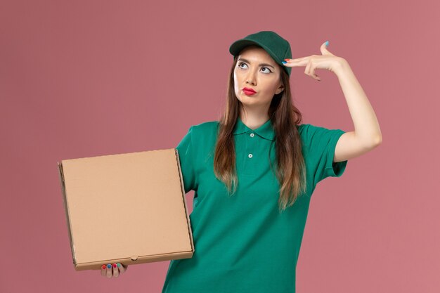 Courier feminino de uniforme verde segurando uma caixa de comida, posando na parede rosa claro, entrega de uniforme de serviço de trabalhador