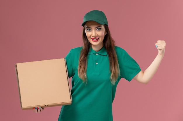 Courier feminino de uniforme verde segurando uma caixa de comida torcendo na parede rosa claro.