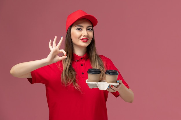 Courier feminino de uniforme vermelho segurando xícaras de café marrons sorrindo sobre fundo rosa claro serviço entrega uniforme trabalhador trabalho feminino empresa