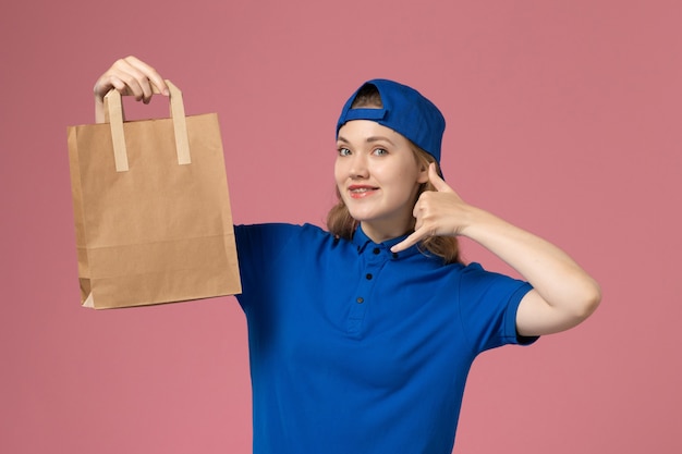 Courier feminino de vista frontal com capa uniforme azul segurando um pacote de entrega de papel na parede rosa, trabalhador de entrega de serviço de trabalho de funcionário
