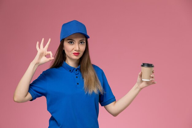 Courier feminino de vista frontal com uniforme azul e capa segurando a xícara de café de entrega na parede rosa mulher garota de trabalho