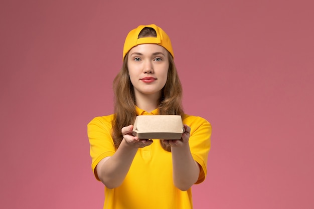 Courier feminino de vista frontal em uniforme amarelo e capa segurando pacote de comida de entrega na parede rosa uniforme de serviço de entrega da empresa trabalho trabalhador garota trabalho