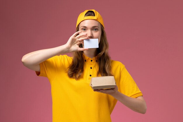 Courier feminino de vista frontal em uniforme amarelo e capa segurando um pequeno pacote de comida de entrega e um cartão de plástico na parede rosa.