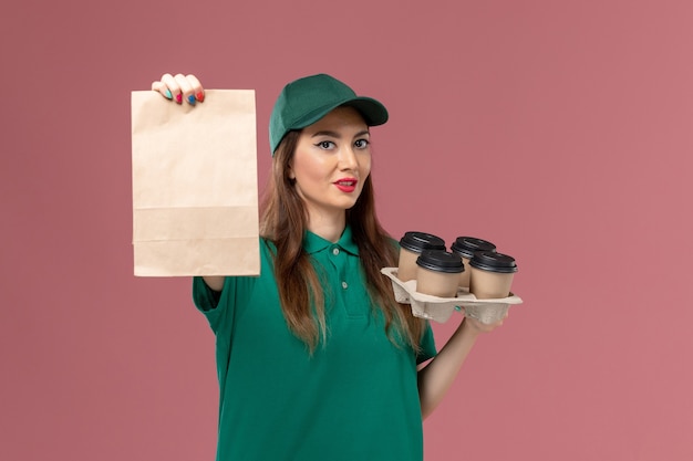 Courier feminino de vista frontal em uniforme verde e capa segurando pacote de comida e entrega de xícaras de café na mesa rosa trabalhador de entrega uniforme de serviço