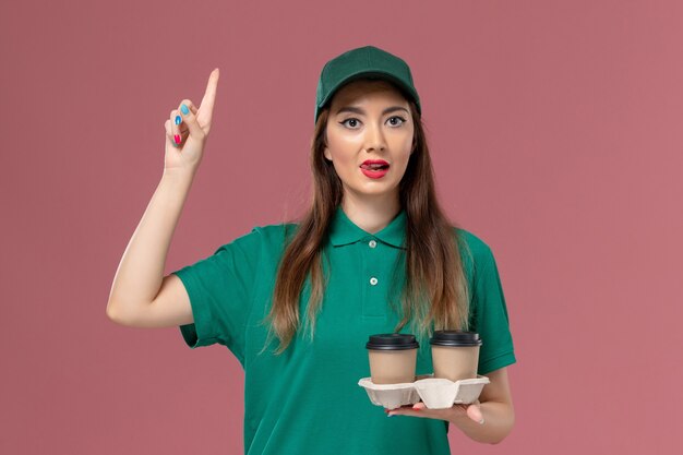 Courier feminino de vista frontal em uniforme verde e capa segurando xícaras de café com dedo levantado na parede rosa empresa serviço trabalho uniforme entrega