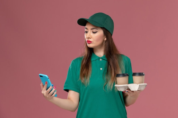 Courier feminino de vista frontal em uniforme verde e capa segurando xícaras de café de entrega e telefone na parede rosa serviço de entrega uniforme de trabalho
