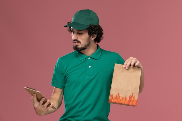 Courier masculino de frente para o uniforme verde e capa segurando o bloco de notas e o pacote de comida no fundo rosa.