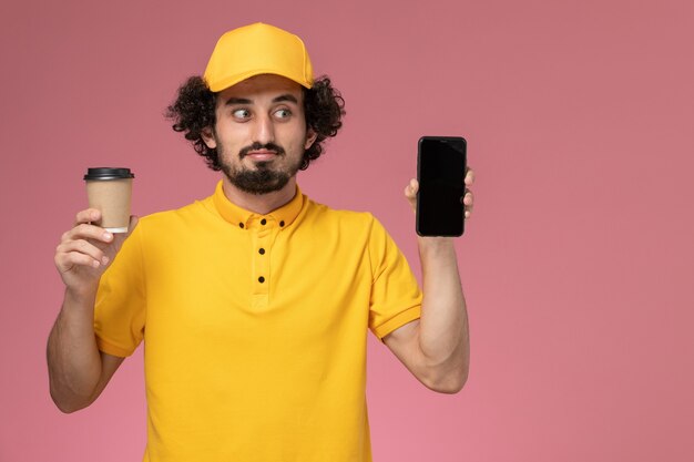 Courier masculino de uniforme amarelo e capa segurando a xícara de café de entrega e o telefone na parede rosa