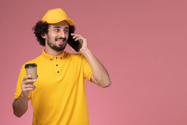 Courier masculino de uniforme amarelo e capa segurando a xícara de café de entrega falando ao telefone na parede rosa