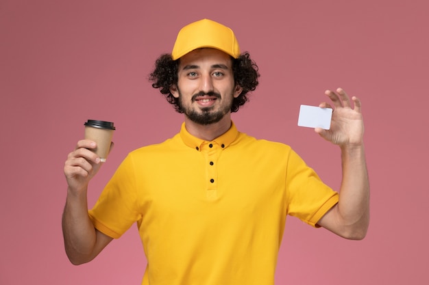 Courier masculino de uniforme amarelo e capa segurando a xícara de café e o cartão de entrega na parede rosa