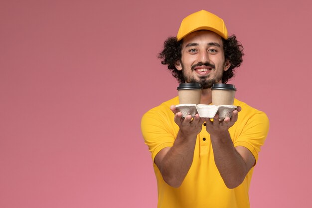 Courier masculino de uniforme amarelo e capa segurando xícaras de café de entrega com um sorriso na parede rosa