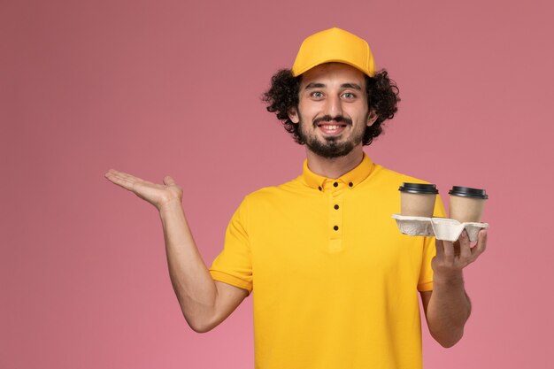 Courier masculino de uniforme amarelo e capa segurando xícaras de café sorrindo na parede rosa