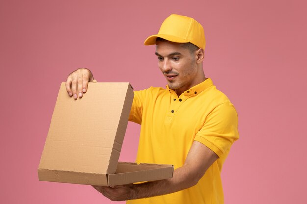 Courier masculino de uniforme amarelo segurando e abrindo a caixa de entrega de comida em fundo rosa