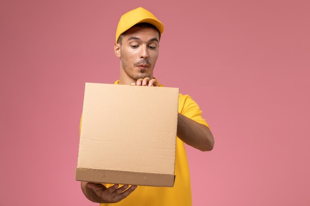 Courier masculino de uniforme amarelo segurando e abrindo a caixa de entrega de comida na mesa rosa