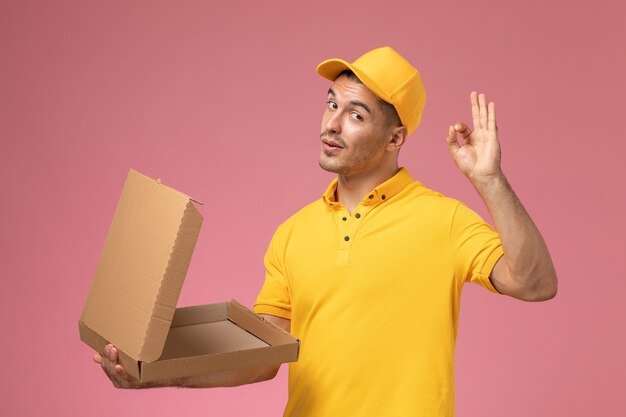 Courier masculino de uniforme amarelo segurando e abrindo a caixa de entrega de comida vazia na mesa rosa