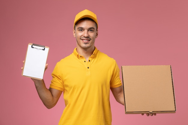 Courier masculino de uniforme amarelo segurando um bloco de notas e uma caixa de entrega de comida com um sorriso na mesa rosa