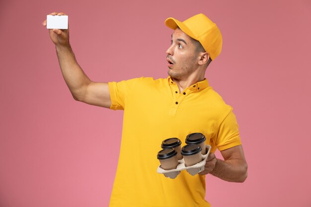 Courier masculino de uniforme amarelo segurando um cartão de plástico e xícaras de café com expressão de surpresa na mesa rosa