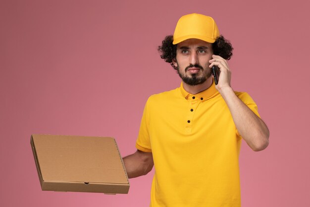 Foto grátis courier masculino de uniforme amarelo segurando uma caixa de entrega de comida e falando ao telefone na parede rosa claro.