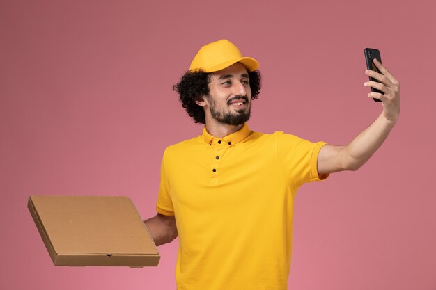 Courier masculino de uniforme amarelo segurando uma caixa de entrega de comida e tirando foto na parede rosa