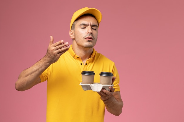 Courier masculino de uniforme amarelo segurando xícaras de café de entrega e cheirando-as em um fundo rosa claro