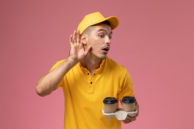 Courier masculino de uniforme amarelo segurando xícaras de café de entrega tentando ouvir na mesa rosa