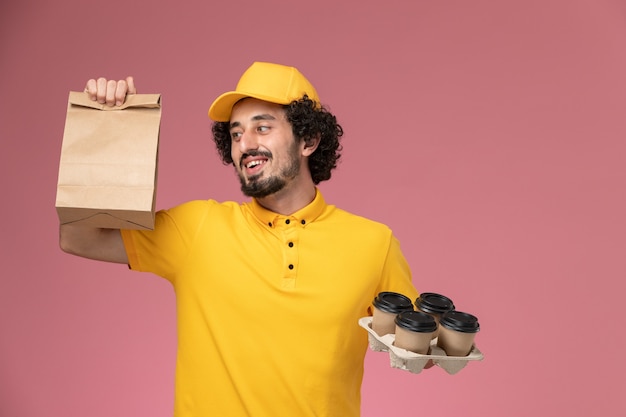 Courier masculino de uniforme amarelo segurando xícaras de café marrons e um pacote de comida na parede rosa